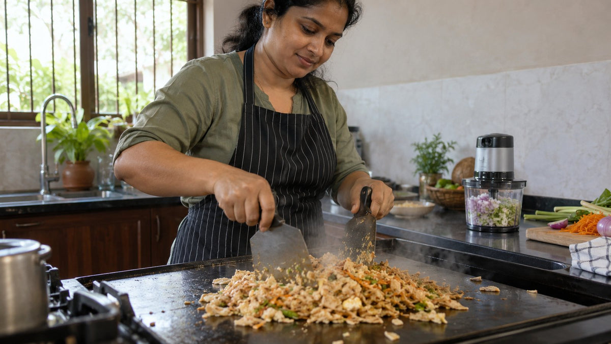 Home cook chopping kottu roti with two blades on a flat griddle, with an electric chopper behind